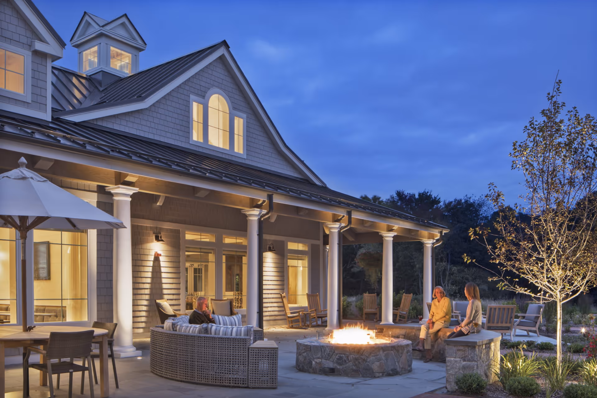 Outdoor patio area at dusk with a fire pit surrounded by seating including a curved sofa and stone benches. Two women are sitting on the stone benches near the fire pit, and another person is seated on the sofa. The building behind has large windows and columns supporting a covered porch with rocking chairs. The area is softly lit with warm lights and there is a tree and some landscaping nearby.