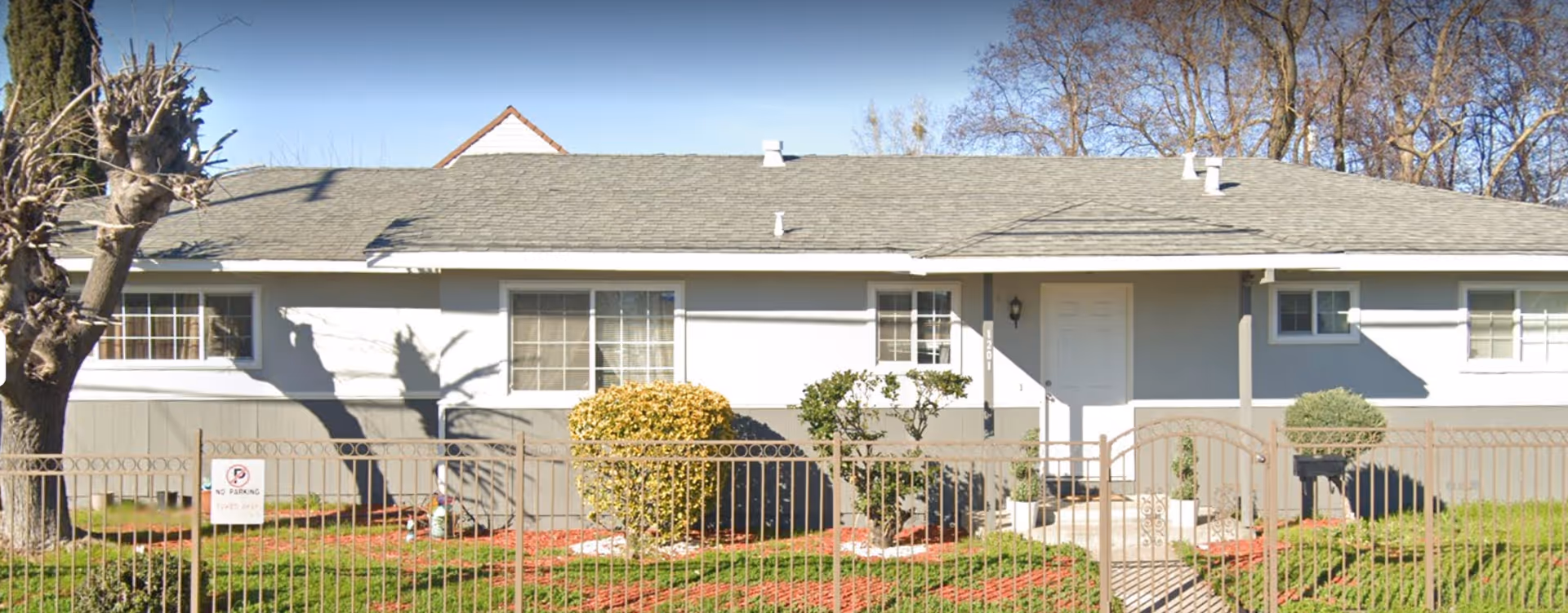 Exterior front view of a single-story residential building with a gray roof and light gray walls. The building has several windows with white frames and a white front door. There is a metal fence with a gate in front, along with some bushes and a tree with pruned branches. A 'No Parking Tow Away Zone' sign is visible on the fence.