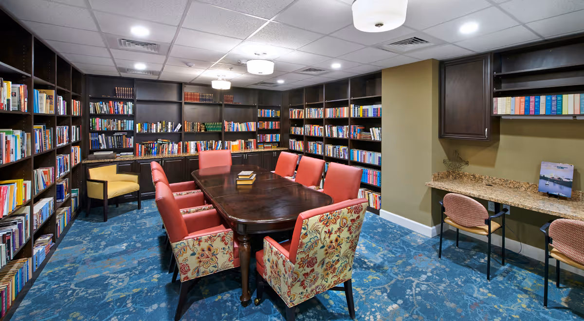 A cozy library room with dark wooden bookshelves filled with books lining the walls. In the center, there is a large wooden table surrounded by eight chairs, six with red upholstery and two with floral patterned upholstery. On the right side, there is a granite countertop with two chairs underneath and a small picture frame on top. The room has a blue patterned carpet and ceiling lights.