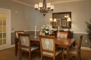A formal dining room with a wooden dining table surrounded by six upholstered chairs. The table is decorated with a floral centerpiece and two lit candles in glass holders. A chandelier with multiple lights hangs above the table. In the background, there is a sideboard with two lamps and a large mirror on the wall. The walls have a patterned wallpaper and there is a door with frosted glass panels on the left side.