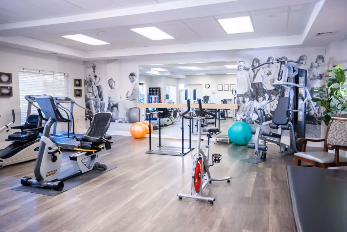 A bright fitness room with exercise equipment including stationary bikes, a treadmill, a multi-function weight machine, and exercise balls. The room has wood flooring, large mirrors on the walls, and black and white photos of people exercising. There is also a chair and a plant in the corner.