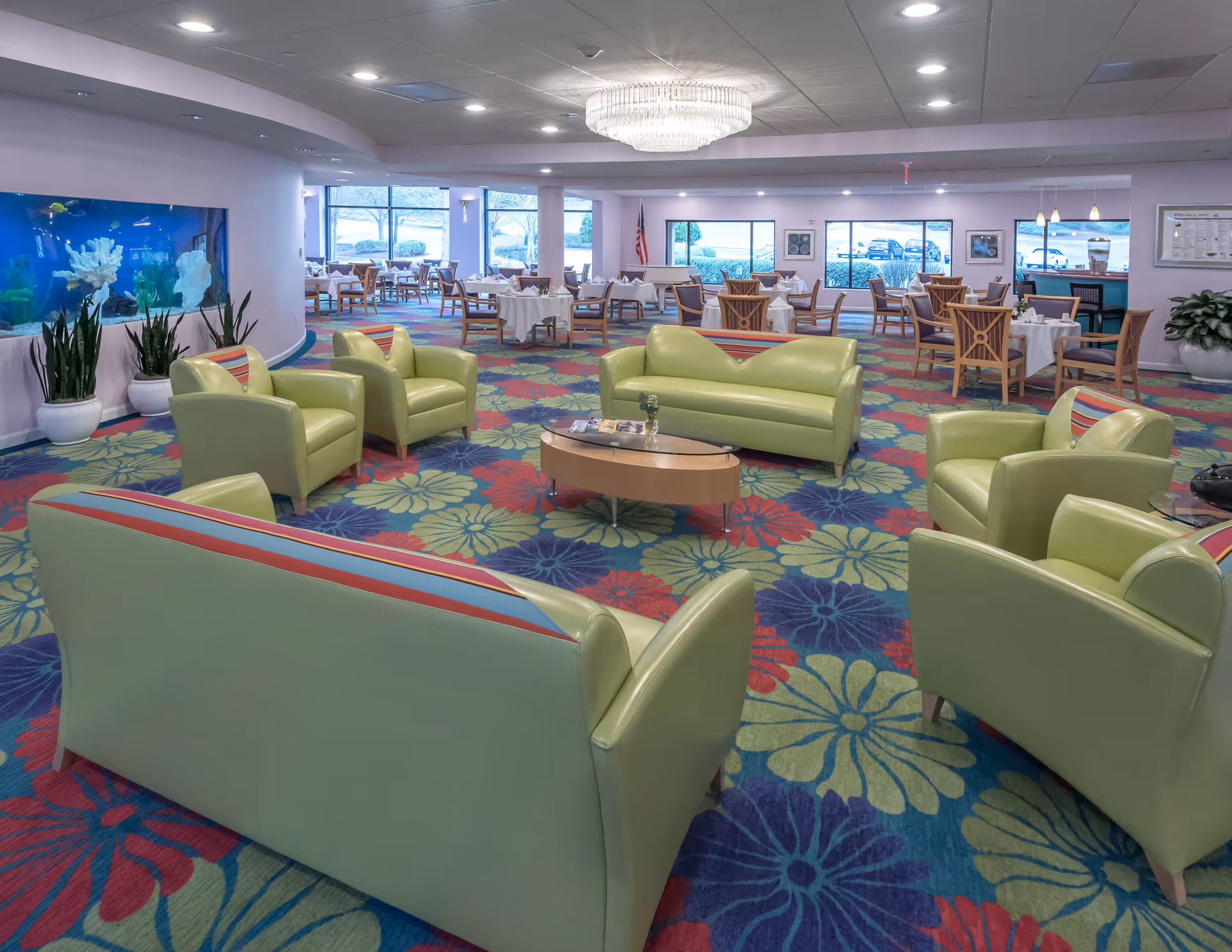 A spacious senior living common area with light green sofas and armchairs arranged around a wooden coffee table. The room features a colorful floral carpet, a large aquarium built into the wall, and several dining tables with chairs set up near large windows letting in natural light. A chandelier hangs from the ceiling, and there are plants placed around the room.