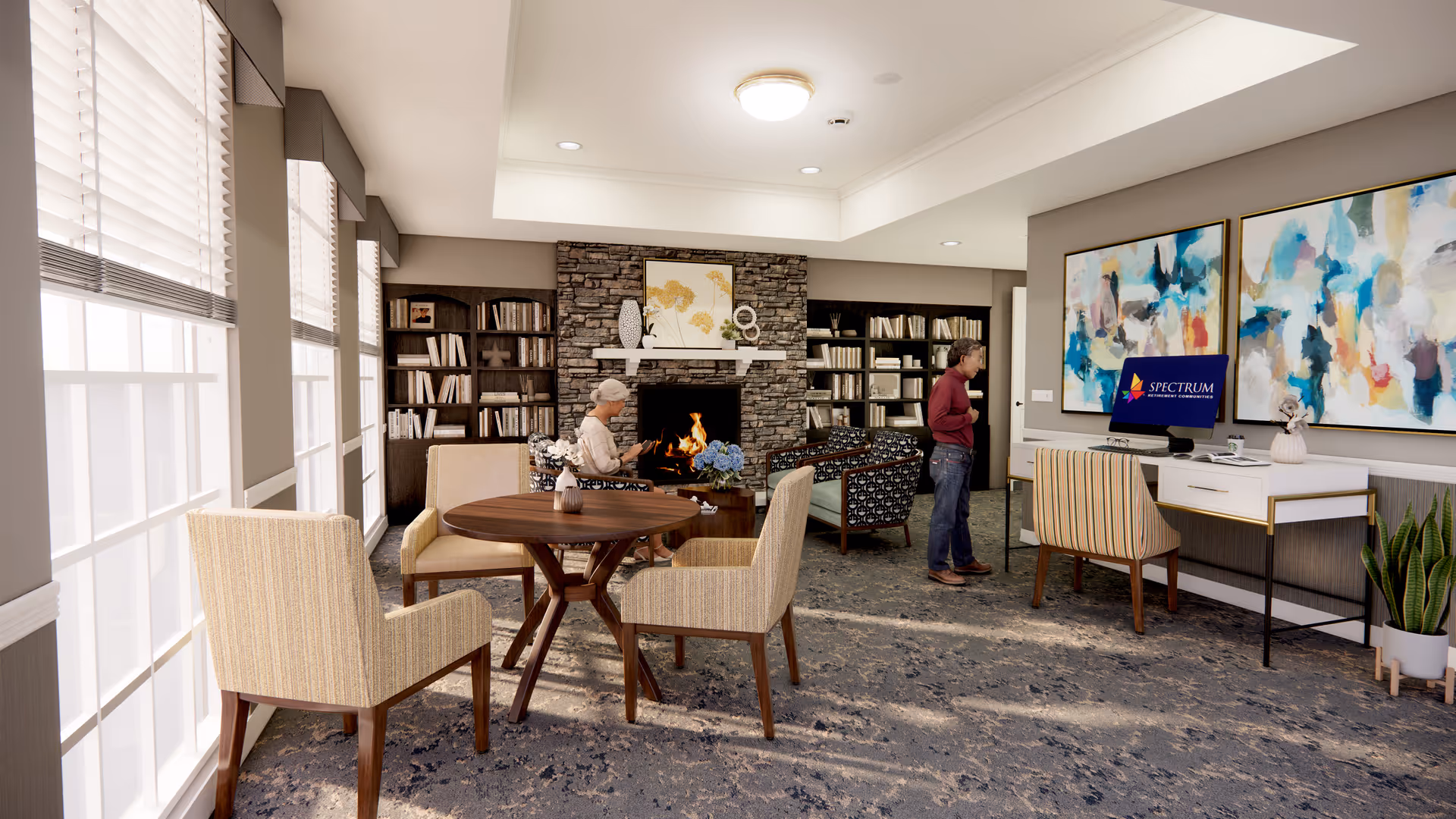 A cozy assisted living facility common area with large windows letting in natural light. There are several beige upholstered chairs around a round wooden table near the windows. In the background, a stone fireplace is lit with a warm fire, flanked by bookshelves filled with books. Two people are present: one seated near the fireplace reading, and another standing near a desk with a computer displaying the Spectrum Retirement Communities logo. The walls are decorated with colorful abstract paintings, and there is a potted plant near the desk.