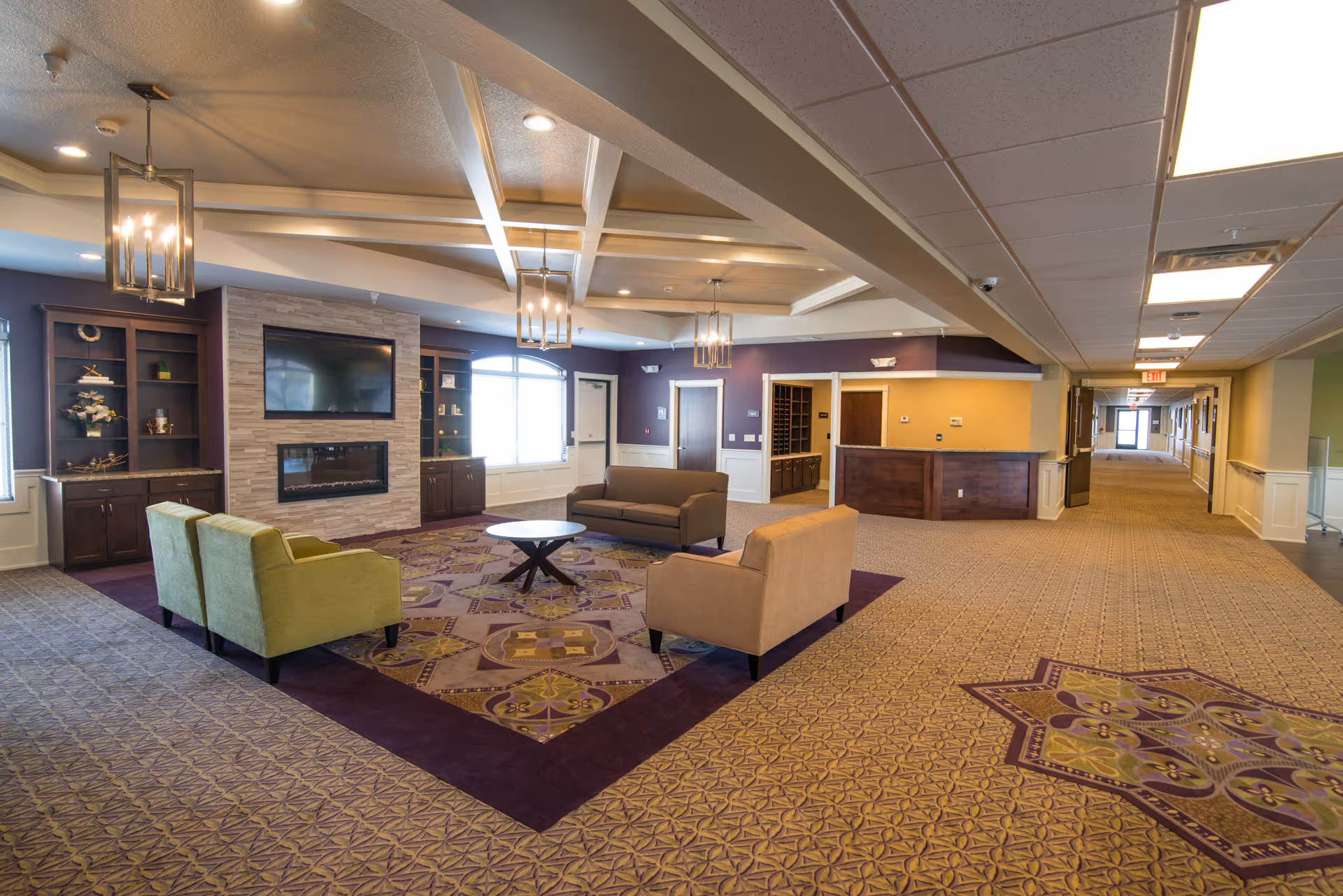 A spacious and well-lit common area in a senior living facility featuring a seating arrangement with two green armchairs, two brown sofas, and a round coffee table on a patterned carpet. The room has a modern fireplace with a mounted flat-screen TV above it, built-in shelves on either side, and decorative ceiling lights. A long hallway extends to the right with doors and an exit sign visible at the end.
