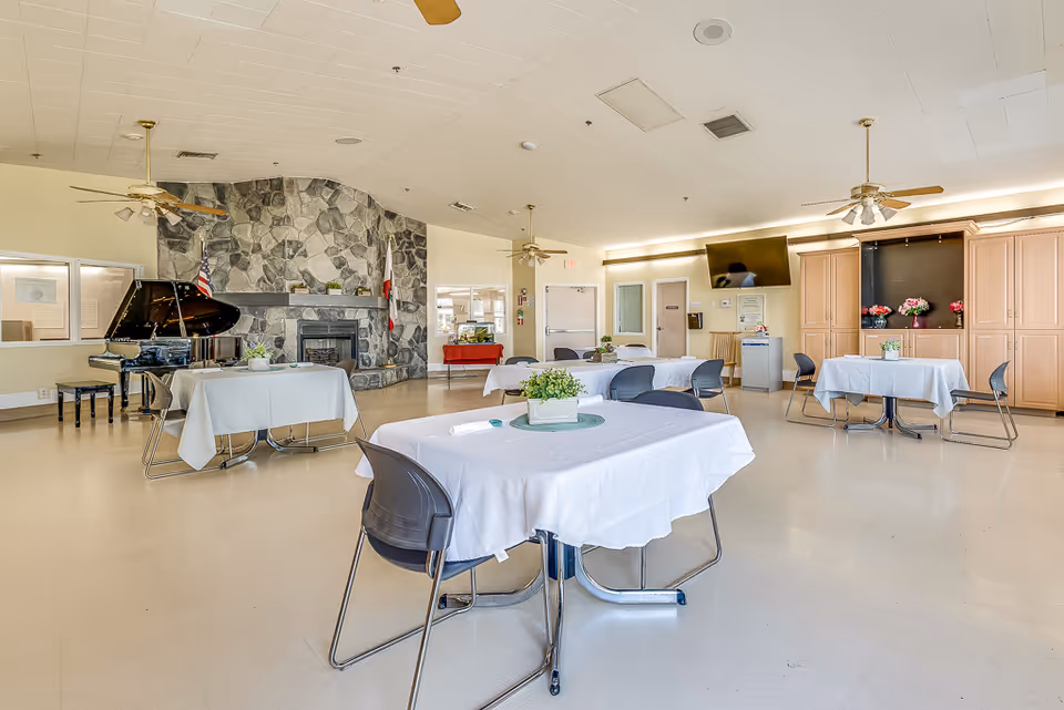Spacious communal dining/activity room with tables covered in white tablecloths, chairs, a grand piano and a stone fireplace.