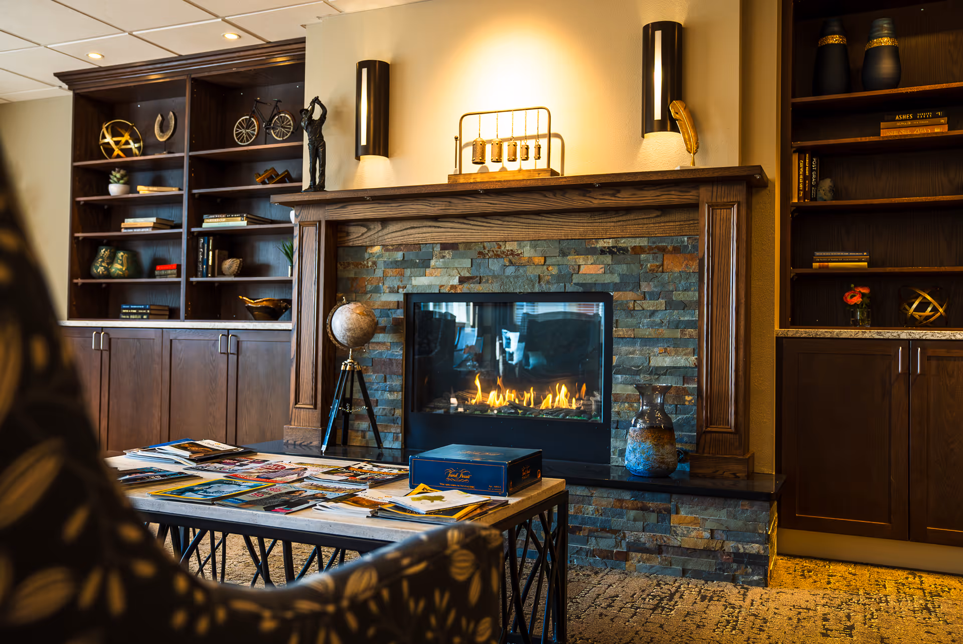 Cozy common room with a lit fireplace set in a stone and wood mantel, built-in bookshelves, and a coffee table covered with magazines.