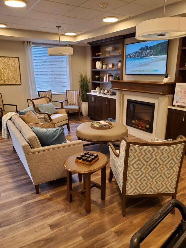 A cozy living room area with a beige sofa, patterned armchairs, a round ottoman, and a small wooden side table with a tic-tac-toe game on it. The room features wood flooring, a fireplace with a TV mounted above it displaying a beach scene, built-in dark wood shelves with decorative items, a large window with white curtains, and a modern ceiling light fixture.