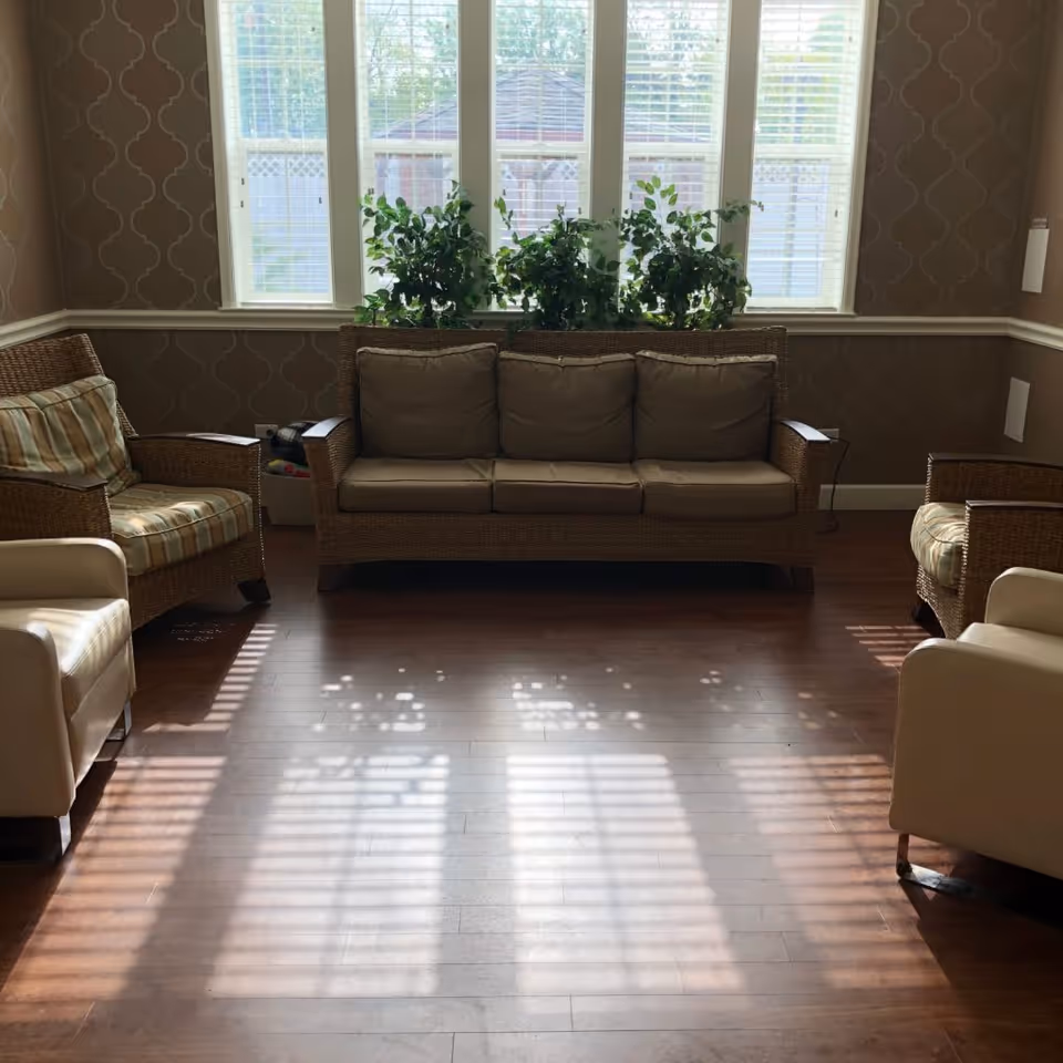 A cozy living room area with a wicker sofa and two wicker armchairs with striped cushions, along with two beige armchairs. The room has wooden flooring, patterned wallpaper, and three tall windows with white blinds letting in sunlight. There are green plants placed on the windowsill behind the sofa.