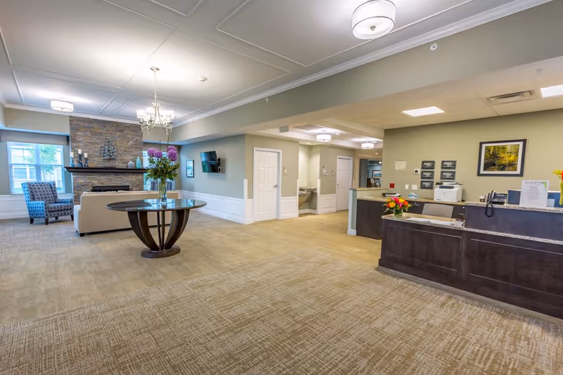 A spacious and well-lit senior living facility lobby area with a round wooden table holding a vase of flowers in the center. To the left, there is a seating area with a white sofa and a patterned armchair in front of a stone fireplace. On the right side, there is a dark wooden reception desk with flowers and office equipment. The walls are painted light green with white trim, and there are ceiling lights and a chandelier providing illumination.