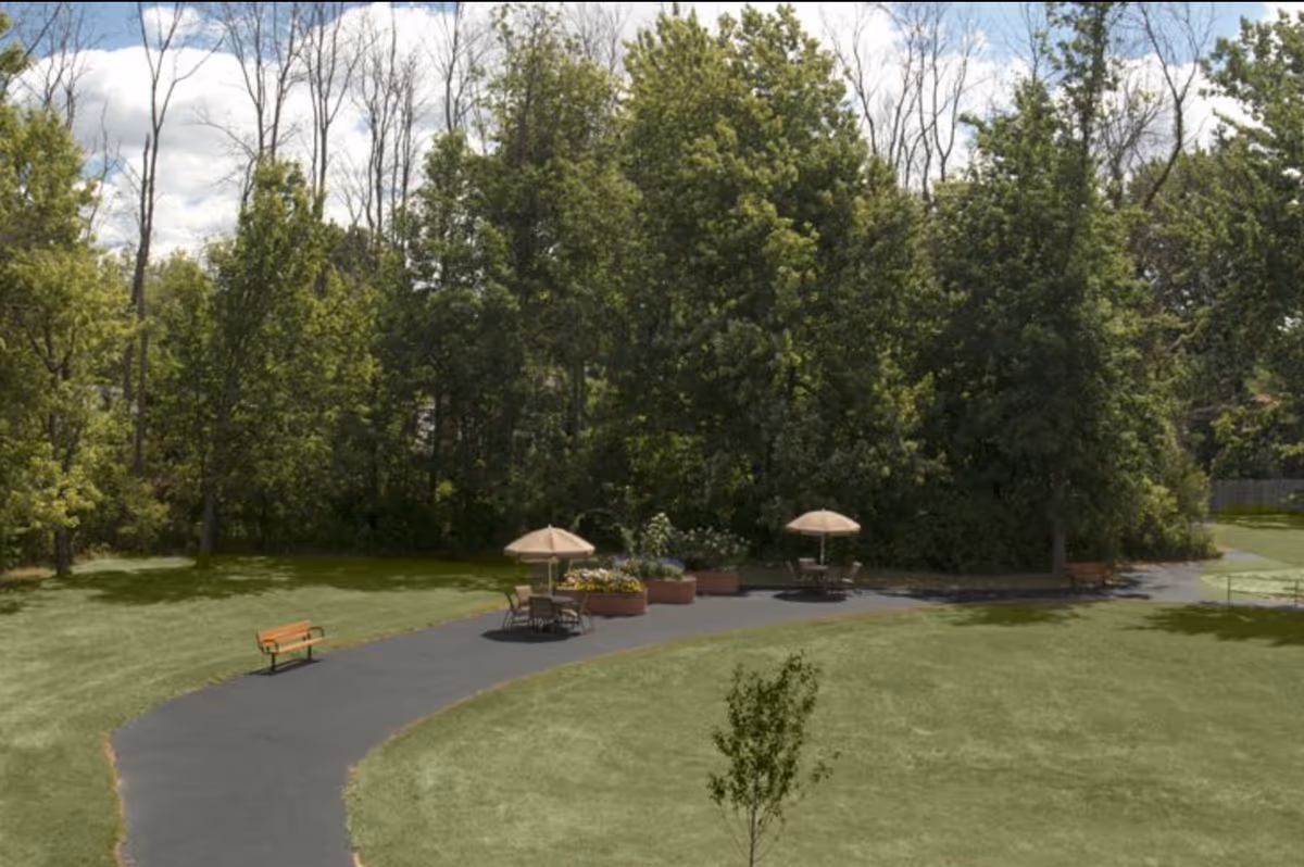 A paved walking path curves through a grassy outdoor area with benches and tables shaded by umbrellas. The background features dense green trees under a partly cloudy sky.