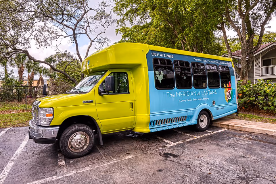 A bright yellow and blue shuttle bus parked in a lot near trees and a building. The bus has signage for The Meridian at Lantana, a luxury senior living community, with a phone number and logo visible on the side.