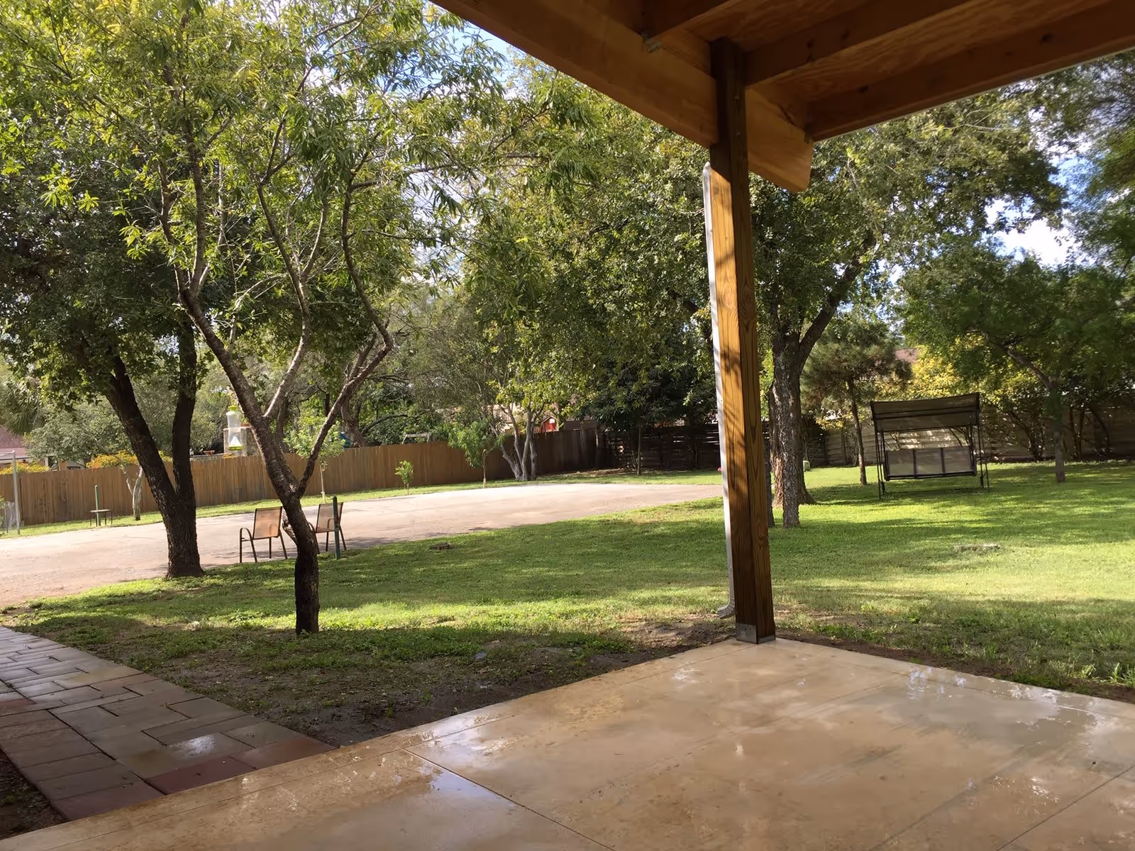 View of a green outdoor area with trees, a wooden fence, two chairs on a paved area, and a covered patio with a wooden post in the foreground.