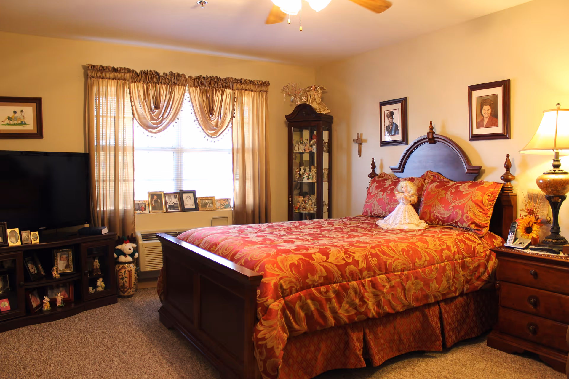 A cozy bedroom with a wooden bed frame and a red and gold patterned bedspread. A doll is placed on the bed. There is a wooden nightstand with a lamp and a telephone on the right side of the bed. A tall glass cabinet filled with decorative items stands in the corner. A window with sheer curtains lets in natural light, and a TV is placed on a wooden stand with framed photos and small decorations. The walls have framed pictures and a cross hanging.