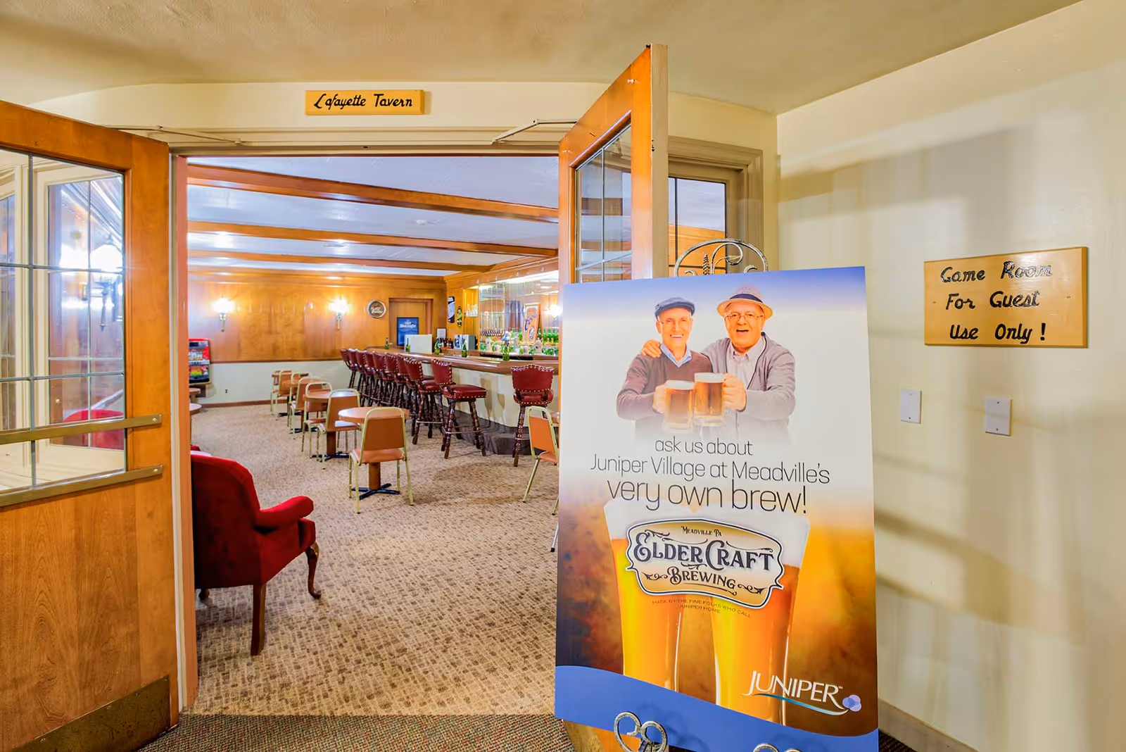 Entrance to Lafayette Tavern featuring an open wooden door with glass panels. Inside, there is a bar with red cushioned bar stools and small tables with chairs. A large sign near the entrance advertises Juniper Village at Meadville's own brew, Elder Craft Brewing, showing two elderly men holding beer mugs. A wall sign indicates the game room is for guest use only.