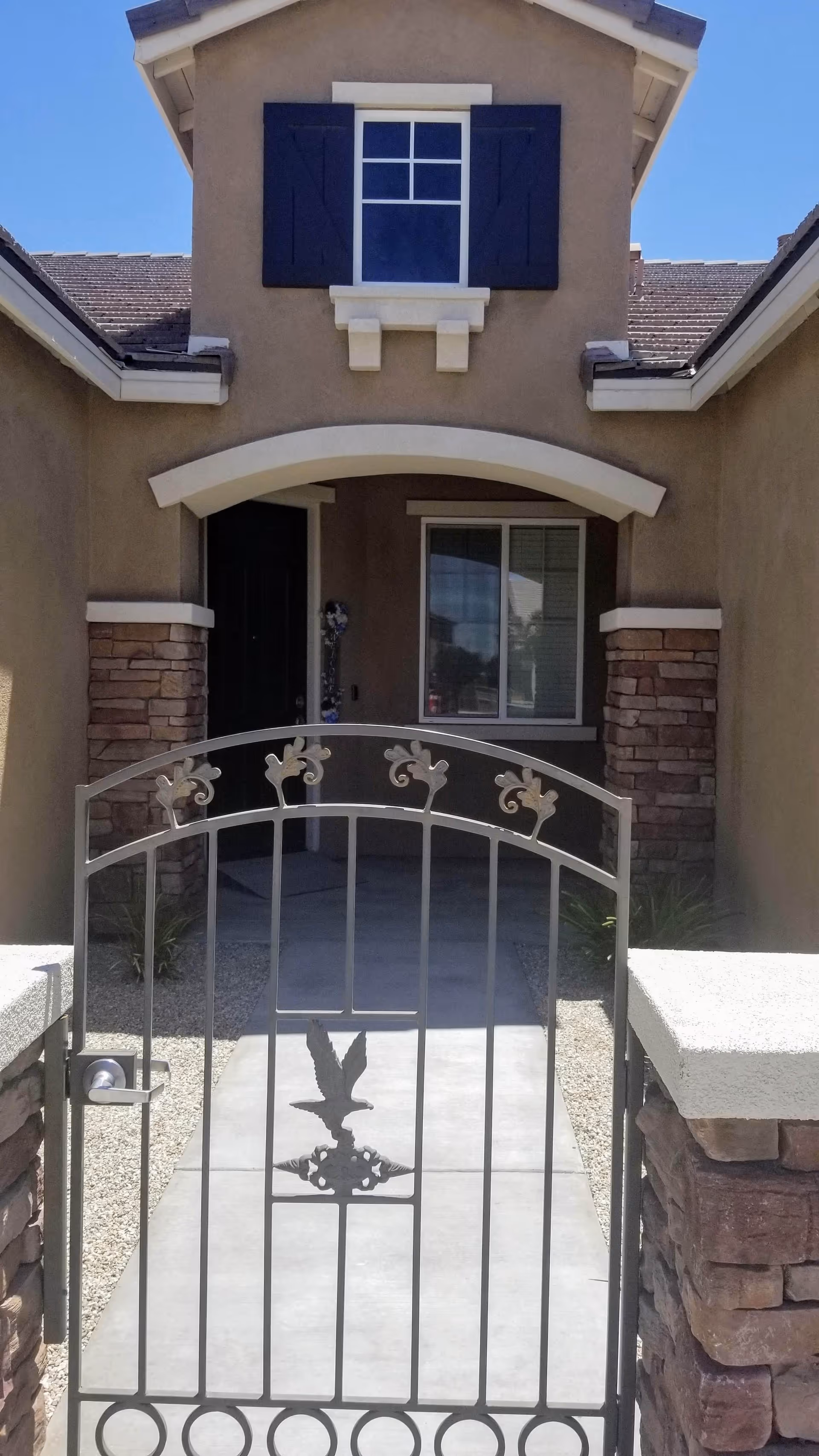 Front entrance of a house with a decorative metal gate featuring an eagle design. The house has a beige exterior with stone accents around the doorway and window. There is a small window with black shutters above the entrance and a concrete pathway leading to the front door.