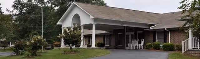 Front exterior of a single-story brick senior living building with a covered entrance, rocking chairs on the porch, driveway and landscaped lawn.
