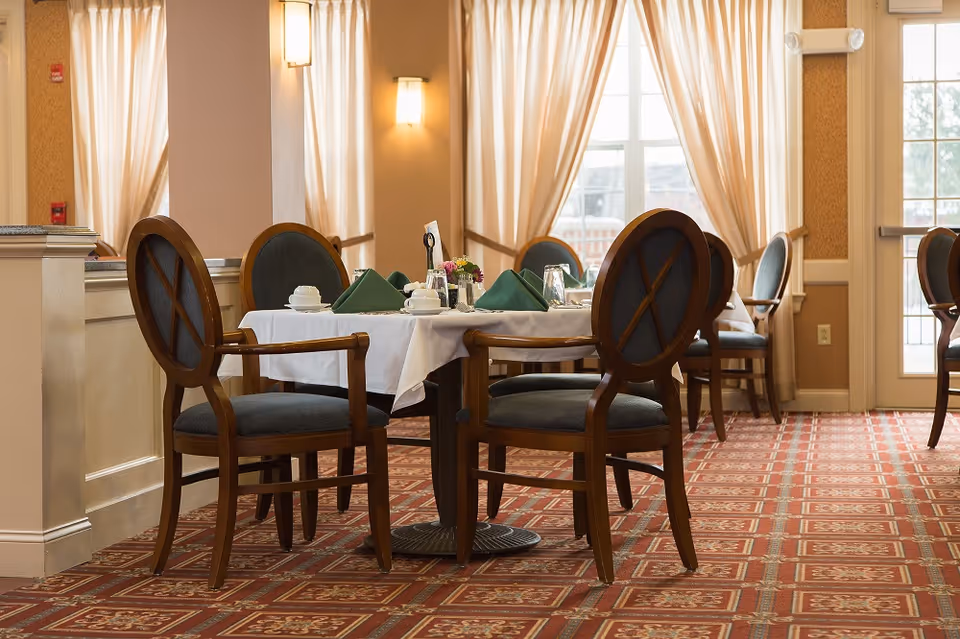 A dining area with a table set for four, featuring wooden chairs with cushioned seats and backs. The table is covered with a white tablecloth and set with green folded napkins, cups, glasses, and a small flower arrangement. The room has large windows with sheer curtains allowing natural light to enter, and the floor is carpeted with a patterned red and beige design.