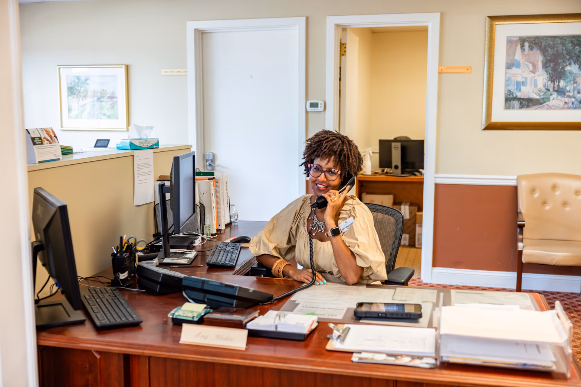 A woman with curly hair and glasses is sitting at a reception desk in an office, talking on the phone. The desk has multiple computer monitors, office supplies, and paperwork. The office has beige walls with white trim, framed artwork, and a beige chair against the wall.