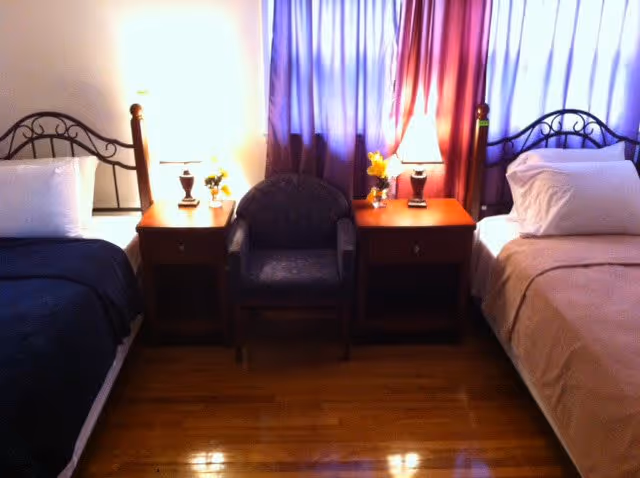 A bedroom with two single beds separated by two wooden nightstands. Each nightstand has a lamp and a small vase with yellow flowers. There is a dark upholstered armchair placed between the nightstands. The beds have metal headboards and are made with white pillows and dark bedspreads. The room has wooden flooring and a window with purple curtains in the background.