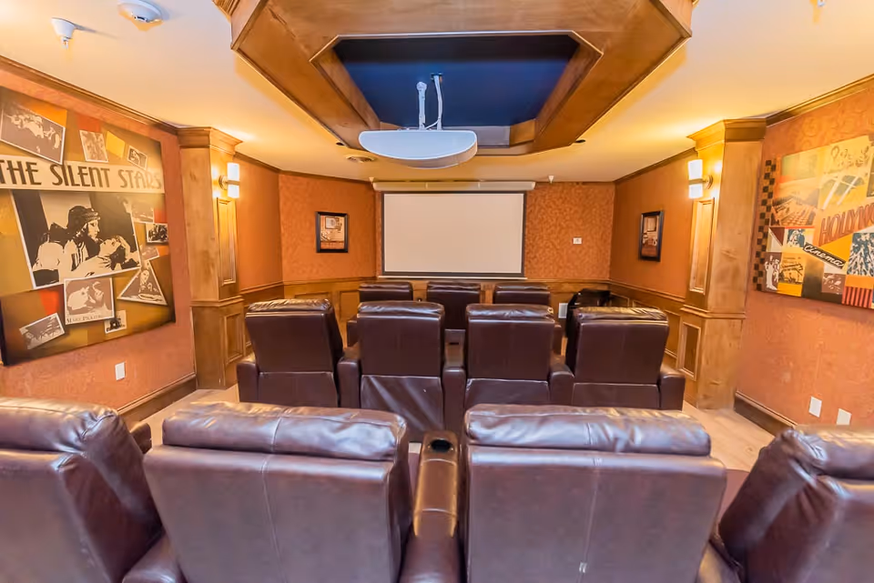 A cozy movie theater room with multiple rows of dark brown leather recliner chairs facing a blank projection screen. The walls are decorated with vintage Hollywood-themed artwork and warm wood paneling, creating an inviting atmosphere.