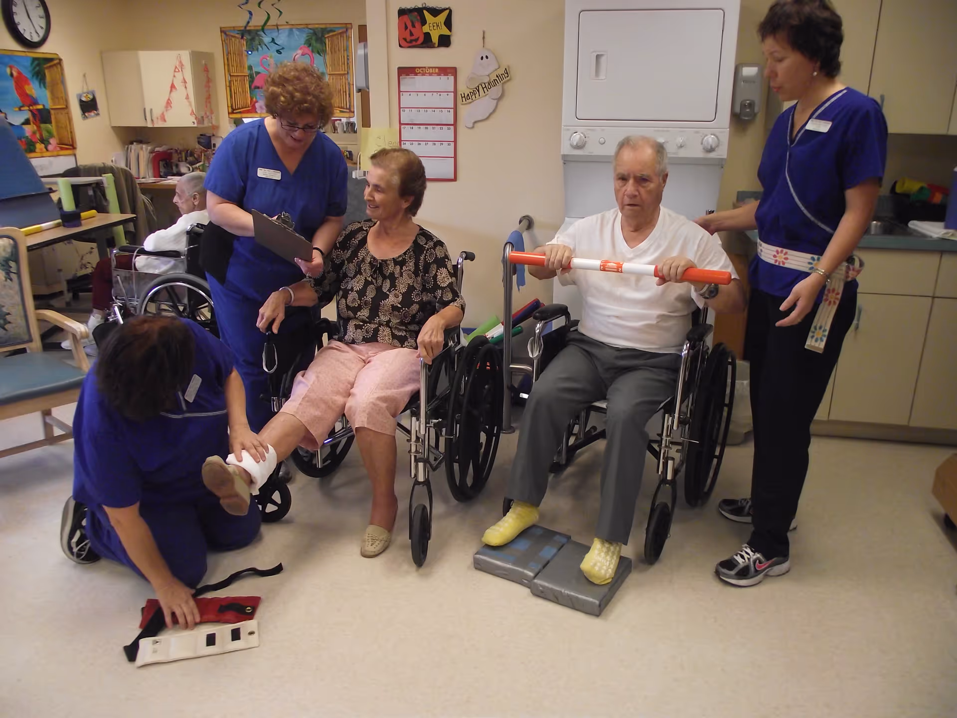 A group of elderly individuals in wheelchairs participating in a physical therapy session with three healthcare workers in blue uniforms assisting them. One elderly woman is having her leg wrapped, while an elderly man is holding a red and white exercise bar and stepping on foam pads. The room has medical and therapy equipment, with decorations on the walls including a calendar and Halloween-themed items.