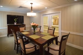 A dining room with a rectangular wooden table set with six chairs. The table has six green placemats and a vase with flowers in the center. A flat-screen TV is mounted on a wooden cabinet against the wall. The room has beige striped wallpaper, a ceiling light fixture, and double doors leading to another area.