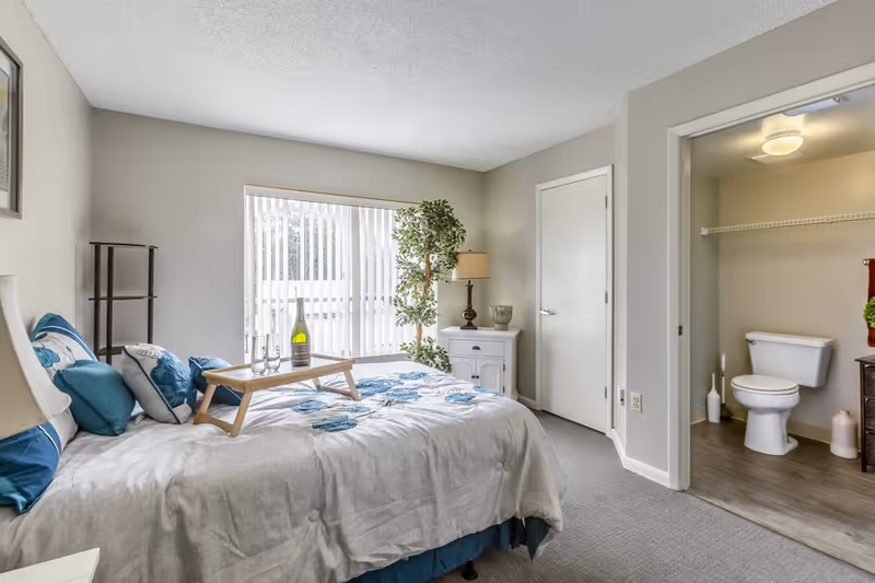 A bedroom with a bed covered in a white and blue floral comforter, several pillows, and a wooden tray holding a bottle of wine and two glasses. There is a large window with vertical blinds, a potted plant, a white nightstand with a lamp and decorative items, and an open door leading to a bathroom with a toilet and shelving.