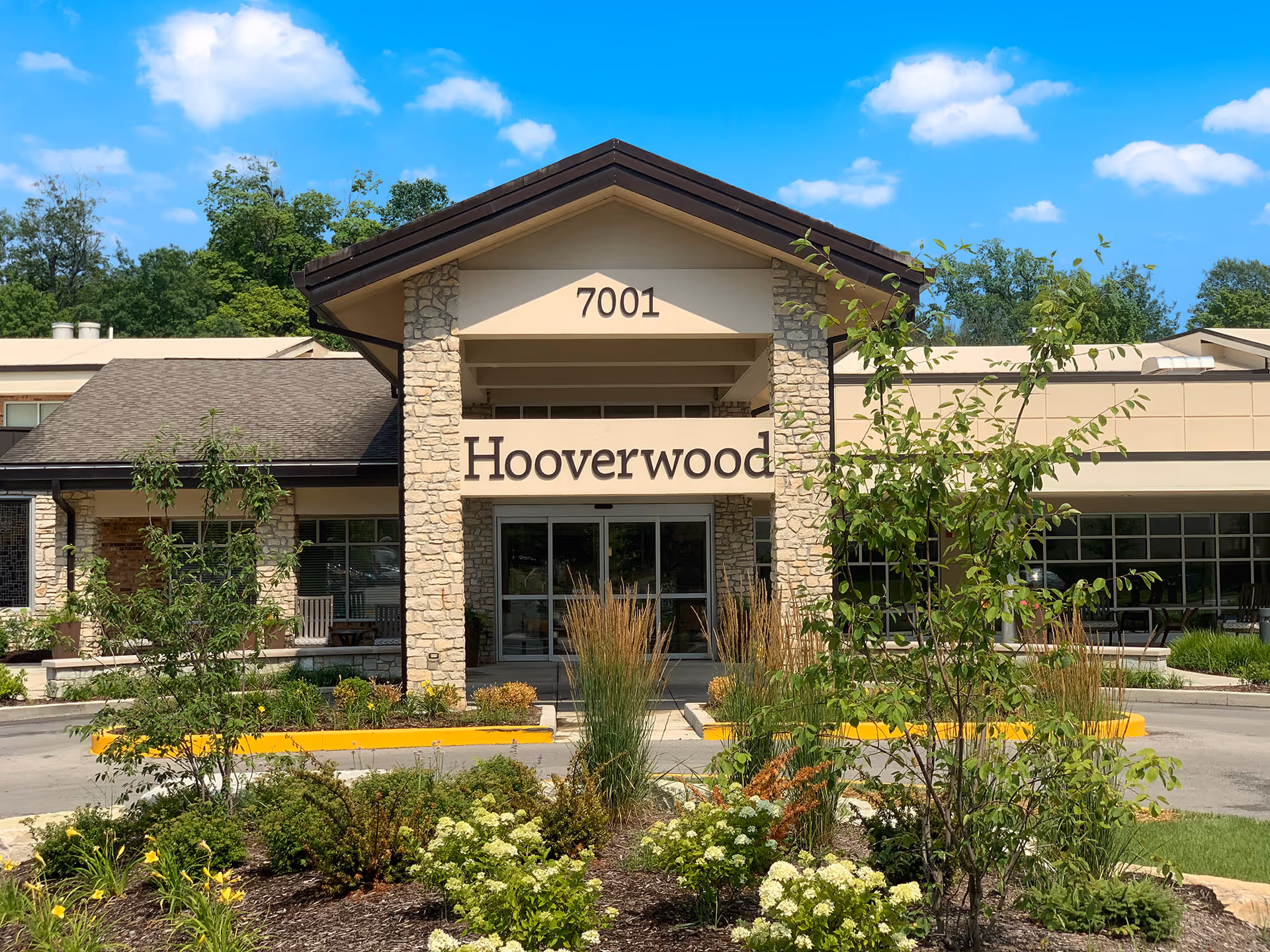 Front exterior view of the Hooverwood Living facility building with stone pillars, a peaked roof, and the address number 7001 above the entrance. There are landscaped plants and shrubs in front of the building under a blue sky with some clouds.