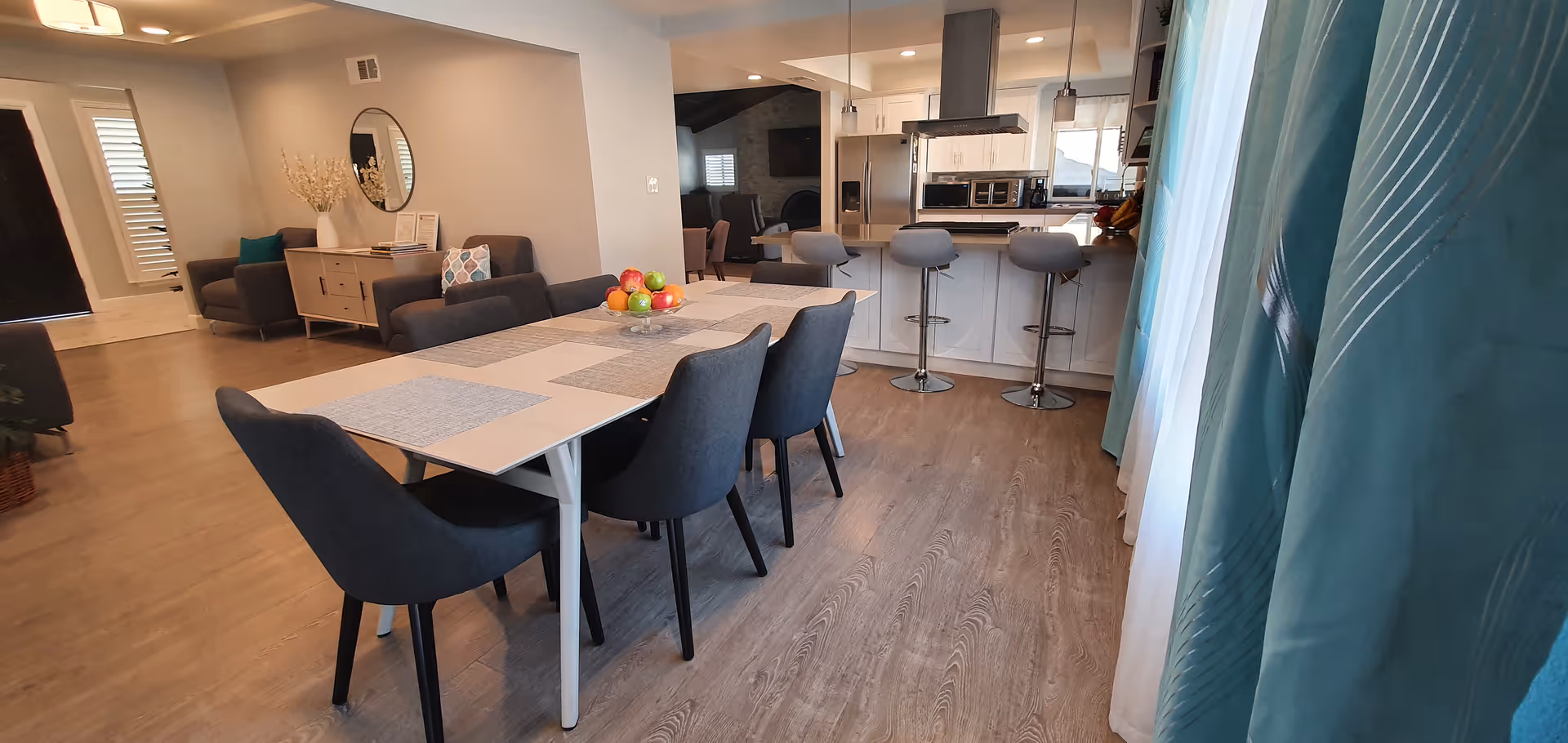 A modern open-concept dining and kitchen area in a senior living facility. The dining area features a rectangular table with six dark upholstered chairs and placemats, with a bowl of fruit as a centerpiece. The kitchen has a white island with three bar stools, stainless steel appliances including a refrigerator and microwave, and light wood flooring throughout. In the background, there is a living area with sofas, a round mirror on the wall, and decorative items on a sideboard.