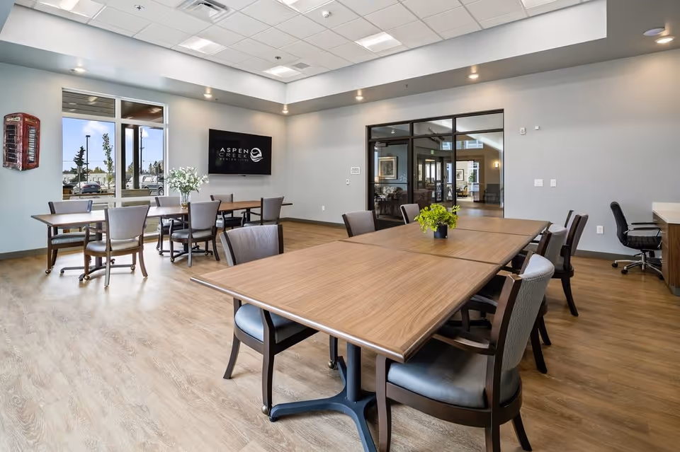 A bright and spacious dining room in Aspen Creek Senior Living featuring wooden tables and cushioned chairs arranged neatly. The room has large windows allowing natural light, a wall-mounted TV displaying the Aspen Creek logo, and modern ceiling lights. There are decorative plants on the tables and a glass door leading to another room.