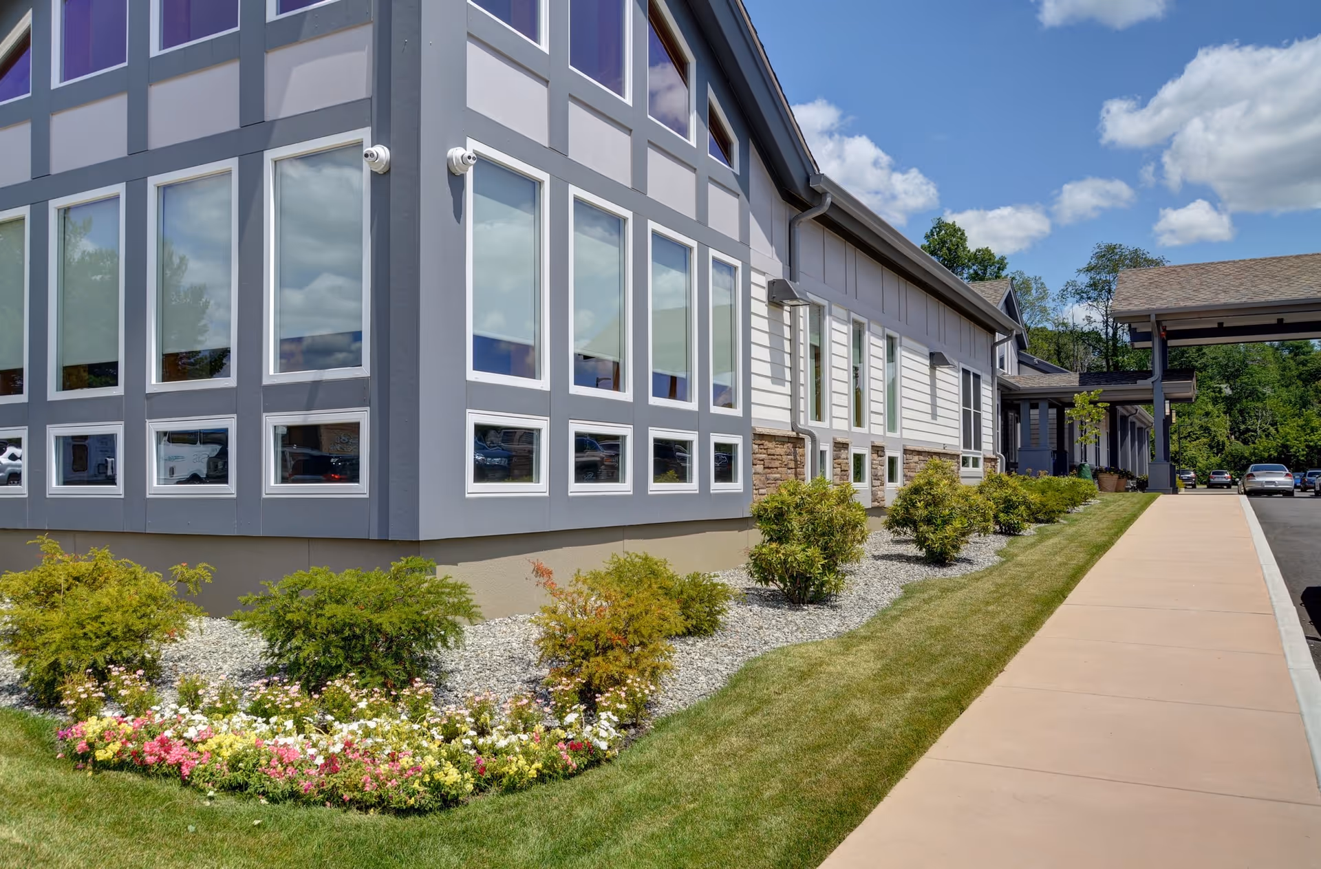 Exterior view of a modern senior living facility building with large windows, landscaped flower beds, shrubs, a sidewalk, and a covered entrance. Cars are parked in the background under a partly cloudy sky.