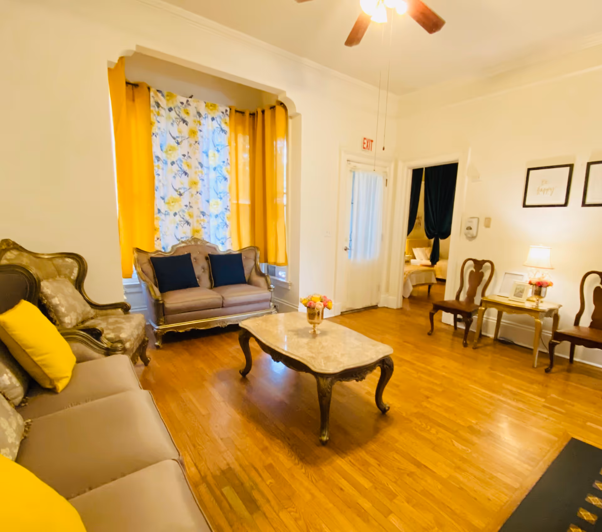 A cozy living room with vintage-style furniture including a sofa, loveseat, and chairs with yellow and dark blue cushions. There is a marble-top coffee table in the center with a flower vase. The room has wooden flooring, yellow and floral curtains on the window, and a ceiling fan with lights. A doorway leads to a bedroom with a bed visible.