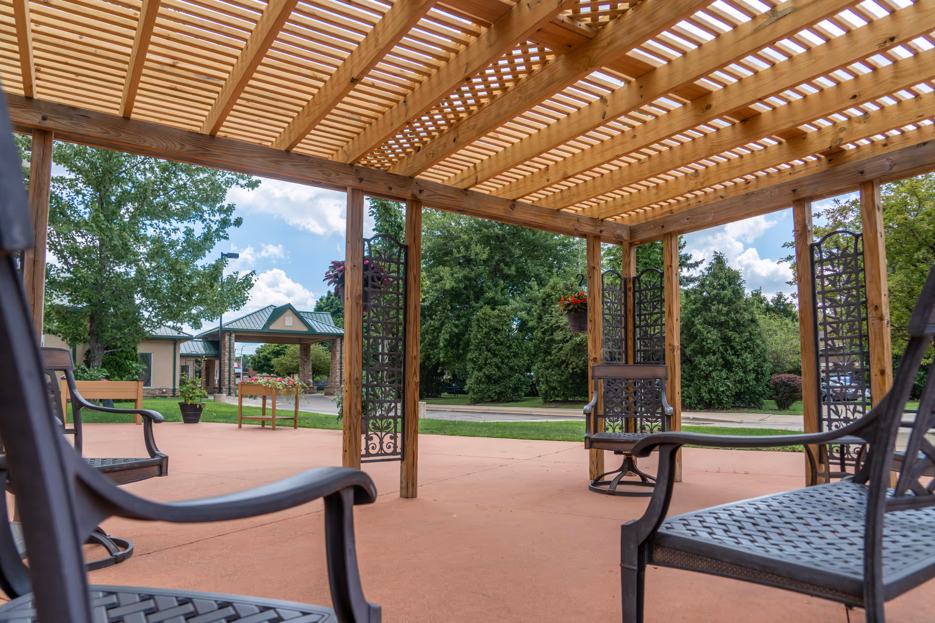 Outdoor seating area under a wooden pergola with metal chairs arranged on a concrete patio. Surrounding the area are green trees and shrubs, with a building entrance visible in the background under a partly cloudy sky.