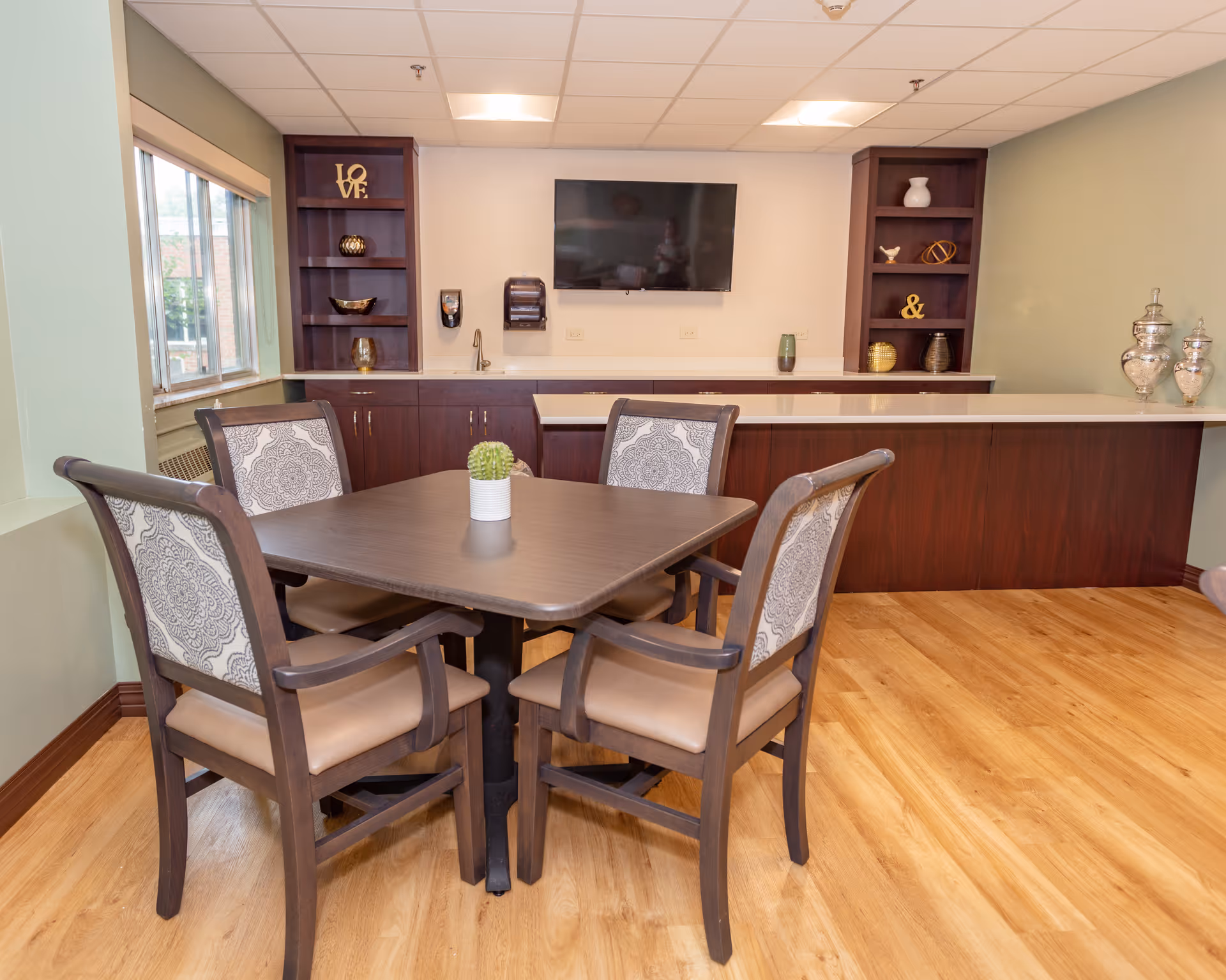 A small dining area with a square wooden table and four cushioned chairs with patterned backs. Behind the table is a counter with dark wood cabinetry and open shelves displaying decorative items. A flat-screen TV is mounted on the wall above the counter. The room has light green walls, a large window, and wood flooring.
