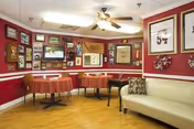 Cozy common room with red walls, framed artwork, small dining tables, a beige sofa, and a ceiling fan.