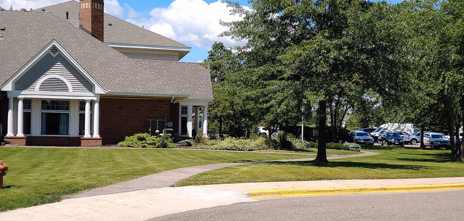 Exterior view of a senior living facility building with a brick and siding facade, a gray shingled roof, and white columns. The building is surrounded by a well-maintained lawn, trees, and shrubs. A sidewalk curves through the grass leading to the building. Several parked cars are visible in the background under the trees.