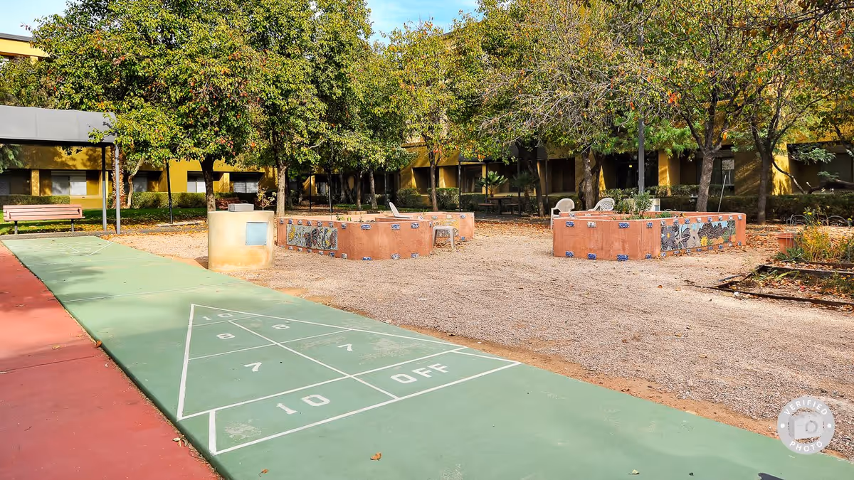 Outdoor area at Broadway Proper Assisted & Senior Living featuring a shuffleboard court, benches, raised garden beds decorated with mosaic tiles, and trees with green and autumn-colored leaves surrounding the space.