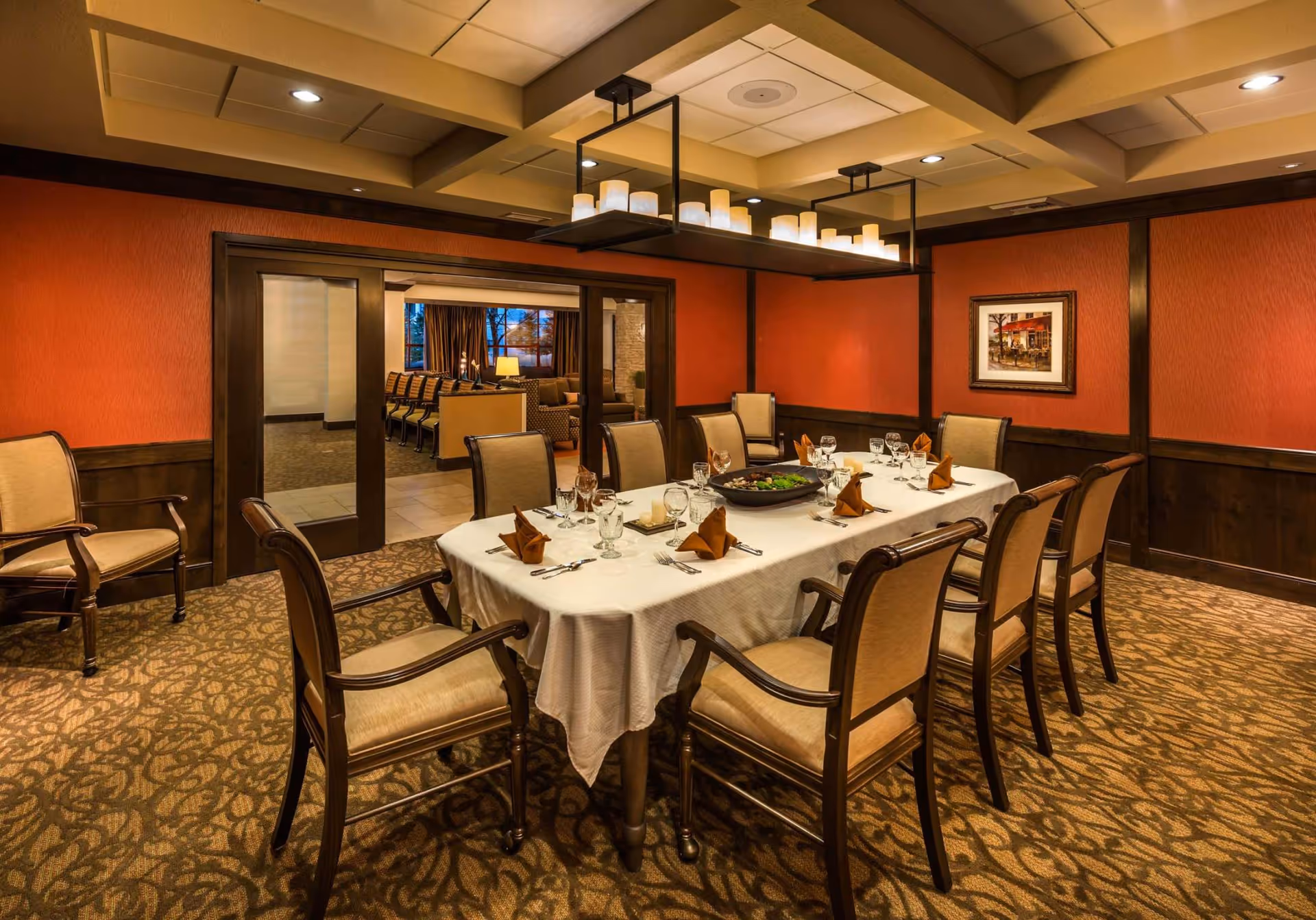 A formal dining room with a long table set for eight, upholstered chairs, warm overhead lighting, and red-walled paneling.