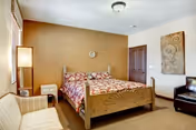 A cozy bedroom with a wooden bed frame featuring a floral bedspread, a beige armchair, a floor lamp, a small nightstand, a dark brown leather chair, and wall decorations including a clock and a patterned artwork.
