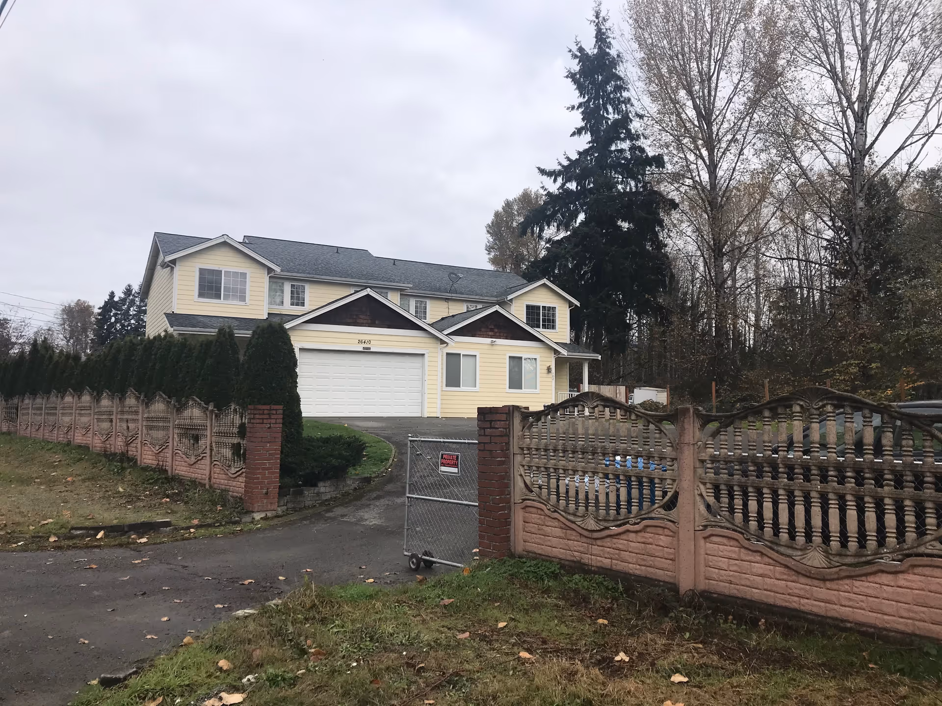 Exterior view of a two-story yellow house with a gray roof, a double garage door, and a driveway leading up to it. The property is enclosed by a decorative fence with brick pillars and a metal gate with a 'Private Property' sign. Tall trees and overcast sky are visible in the background.