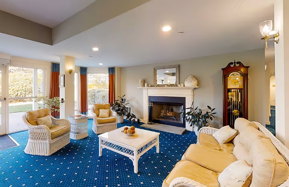 A cozy living room with a blue carpet featuring a white dotted pattern, beige cushioned wicker furniture including a sofa and two armchairs, a white coffee table with a bowl of decorative items, a fireplace with a white mantel, a large mirror above the fireplace, potted plants on either side of the fireplace, tall windows with orange and blue curtains, and a wooden grandfather clock in the corner.