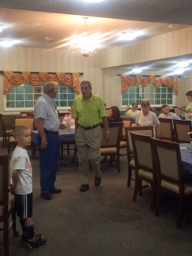 A dining room in a senior living facility with several elderly people seated at tables covered with blue tablecloths. Two elderly men are standing and talking near the center, and a young boy stands nearby. The room has beige walls, large windows with orange curtains, and ceiling lights.