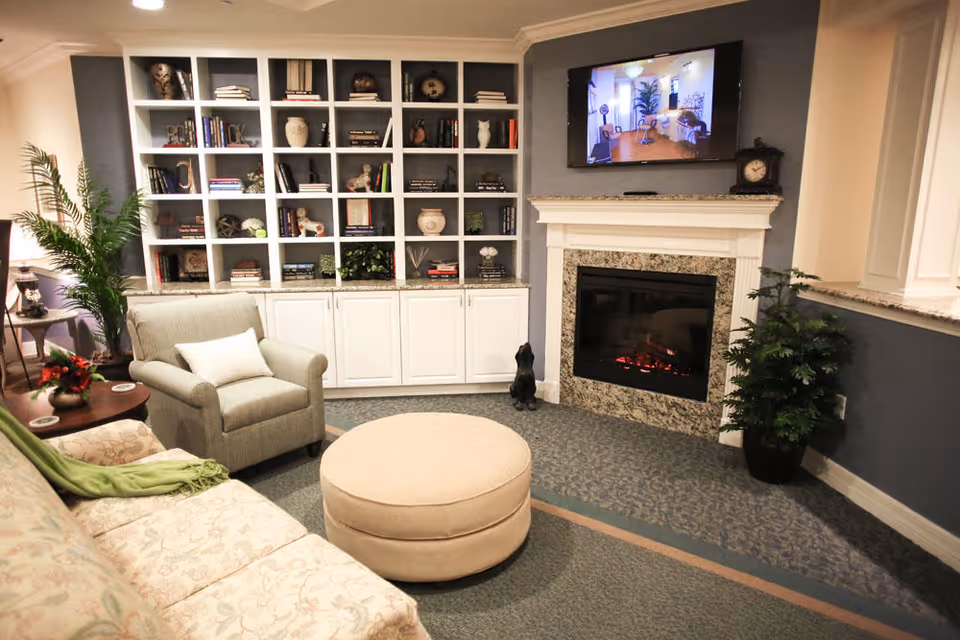 Cozy living room with a floral patterned sofa, a cushioned armchair with a white pillow, and a round beige ottoman. There is a built-in white bookshelf filled with books and decorative items against the wall. A fireplace with a granite surround is below a mounted flat-screen TV displaying an image of another room. Two potted plants add greenery to the space.