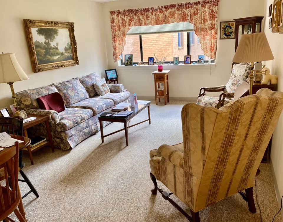 A cozy living room with floral patterned sofa and armchair, a wooden coffee table with books and papers, a side table with a lamp, a large window with floral curtains, and framed artwork on the walls.
