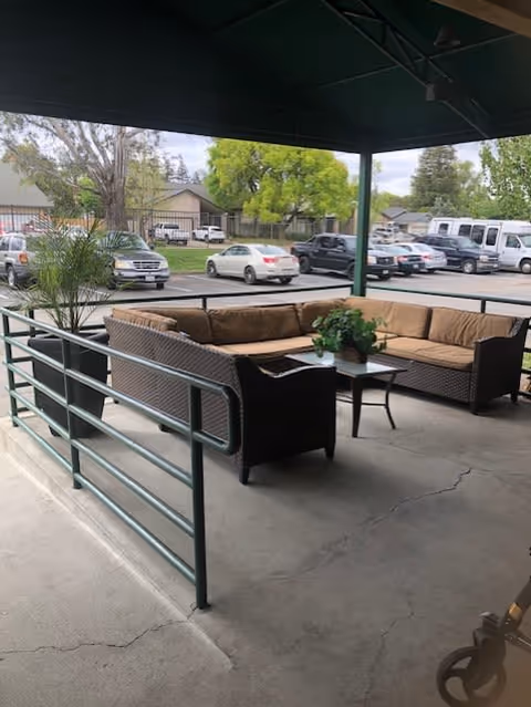 Covered outdoor seating area with a brown cushioned sectional sofa and a small table with a potted plant. The area is surrounded by a metal railing and overlooks a parking lot with several parked cars and trees in the background.