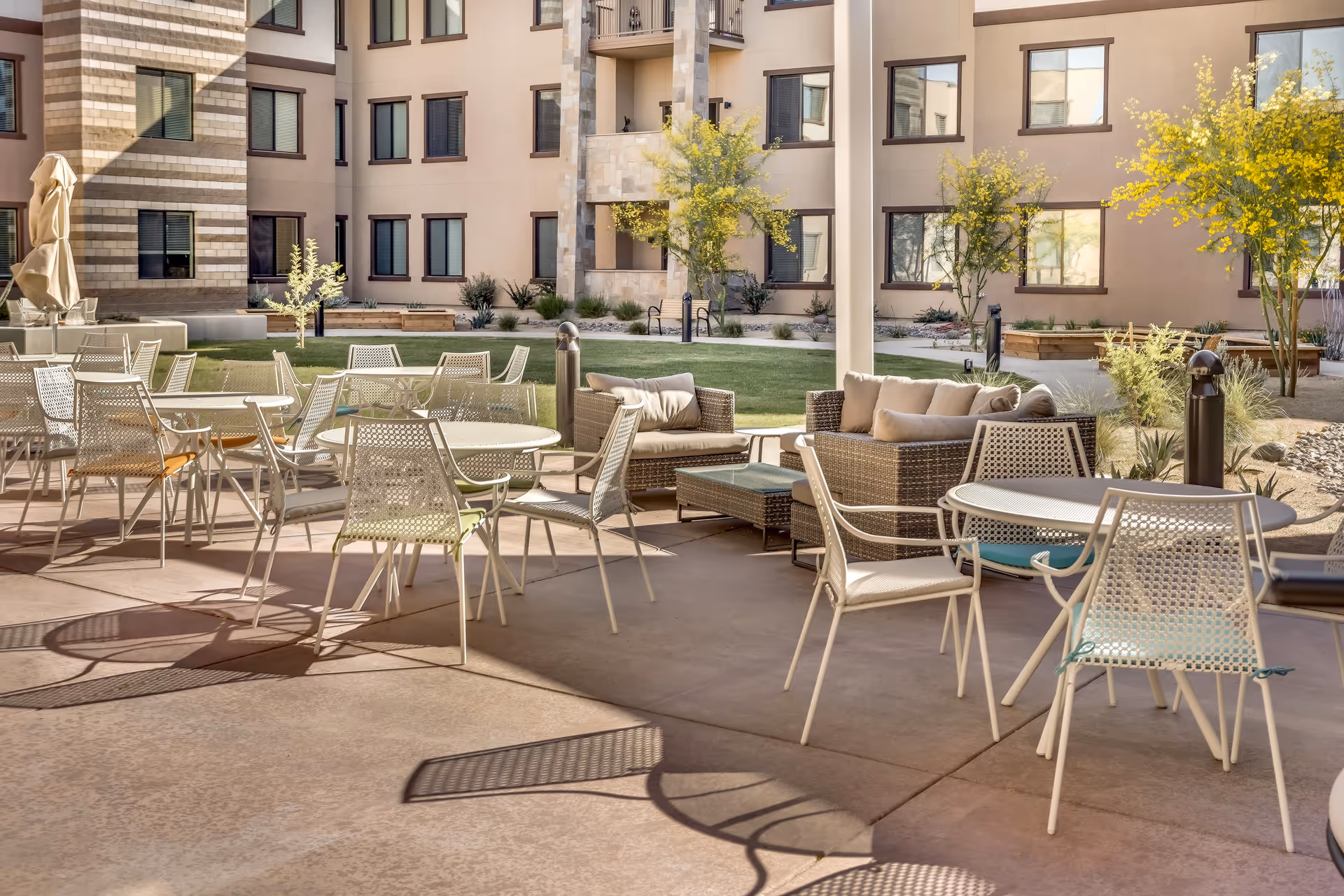 Outdoor patio area at Legacy Village of Salt River with multiple round tables and chairs, cushioned wicker sofas, green lawn, small trees, and a multi-story building in the background.