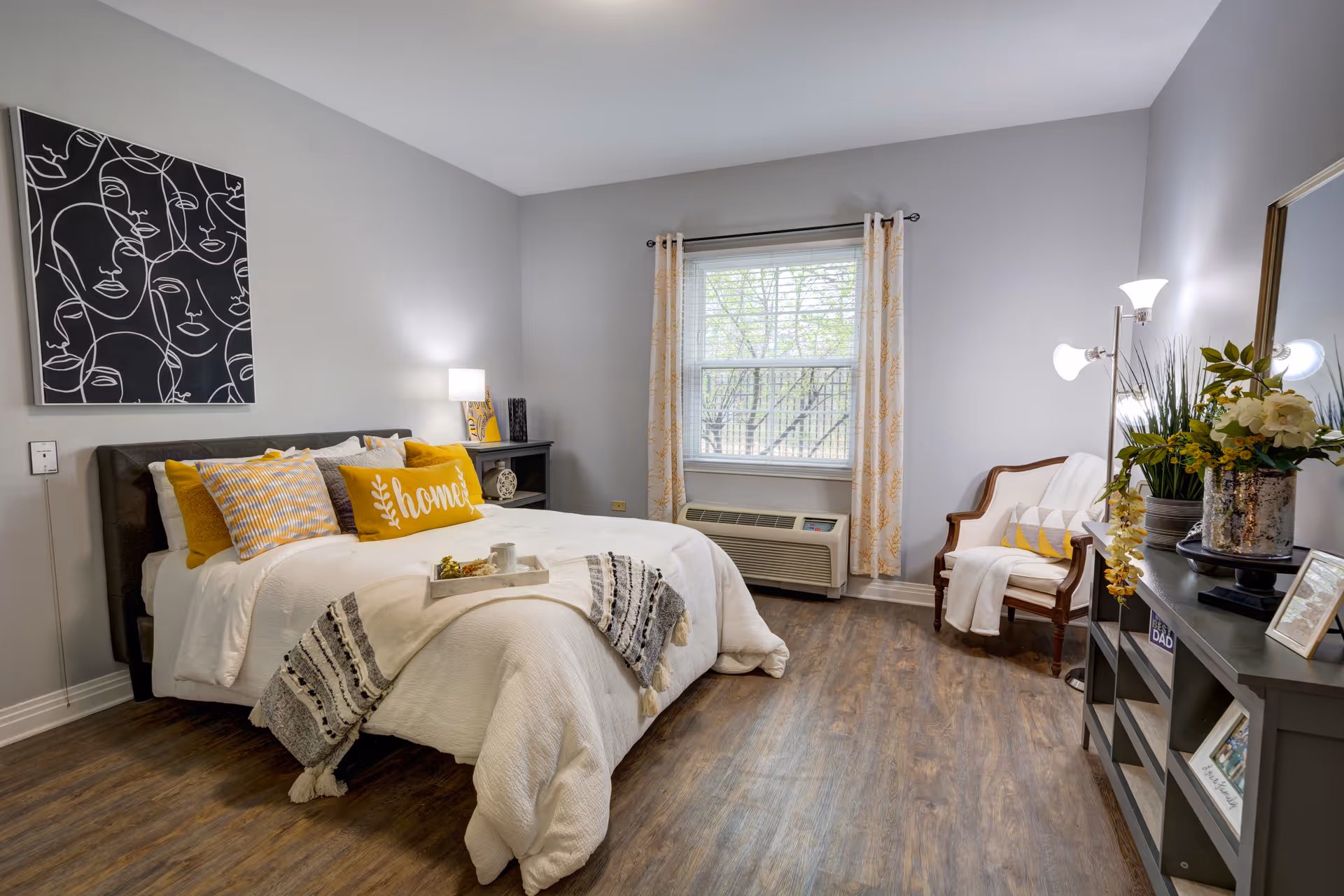 A cozy bedroom with a neatly made bed featuring white bedding and yellow accent pillows, including one with the word 'home'. The room has light gray walls, a window with patterned curtains, a wooden floor, a small black nightstand with a lamp, a wooden chair with a white throw, and a dark dresser with a mirror and decorative plants and picture frames.