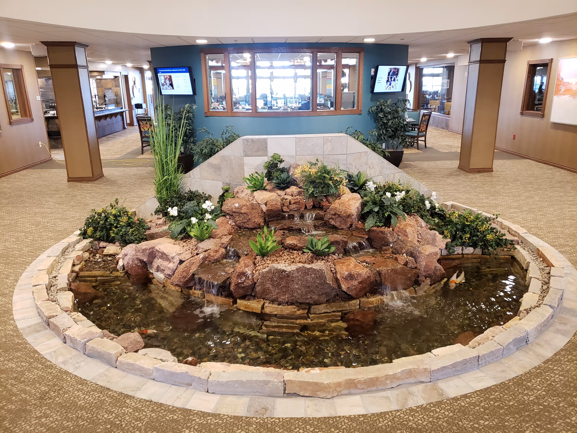 Indoor water feature with rocks, small waterfalls, and plants in the center of a carpeted room with wooden pillars and windows looking into another room with tables and chairs.