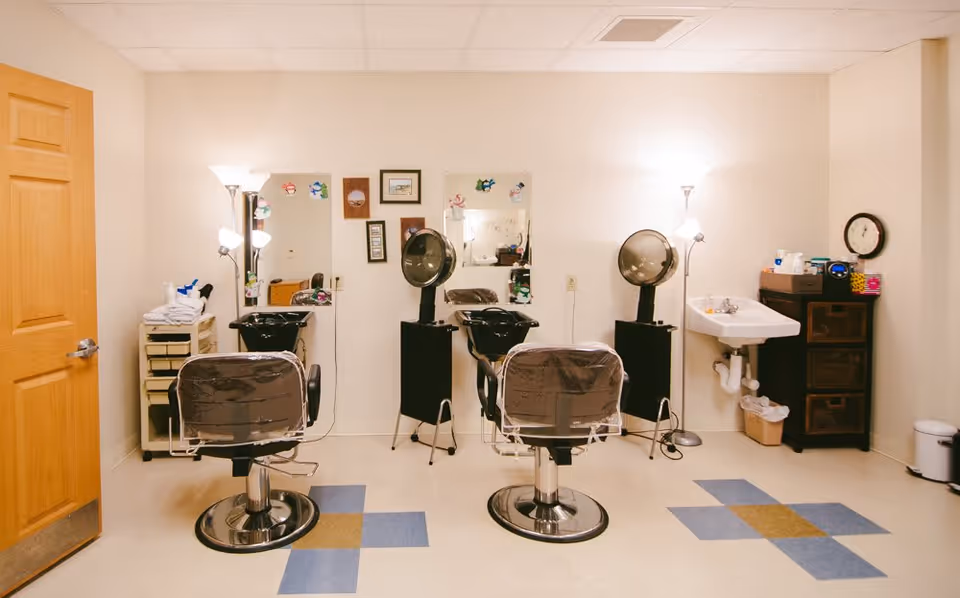 Interior view of a hair salon area in a senior living facility with two salon chairs facing mirrors, hair dryers, a sink, and storage cabinets. The room has a tiled floor with blue and brown patterns and is softly lit by floor lamps.
