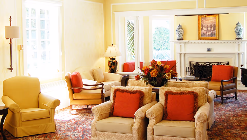 A bright and cozy living room with multiple upholstered armchairs and sofas arranged around a central coffee table with a floral arrangement. The room features large windows letting in natural light, a fireplace with decorative vases and a framed painting above it, and a patterned carpet covering the floor.